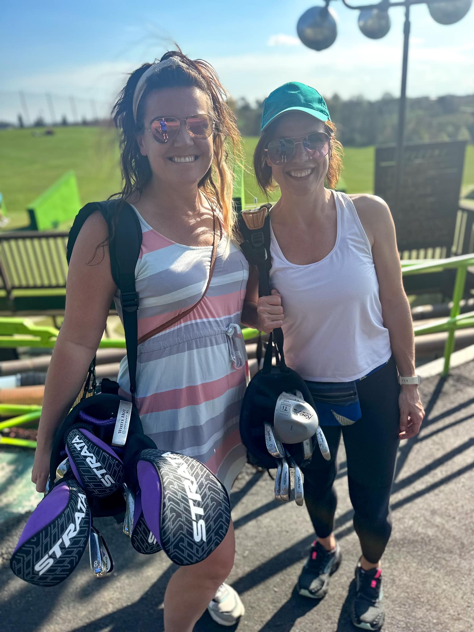 Two women standing at the driving range holding bags of golf clubs over their shoulders